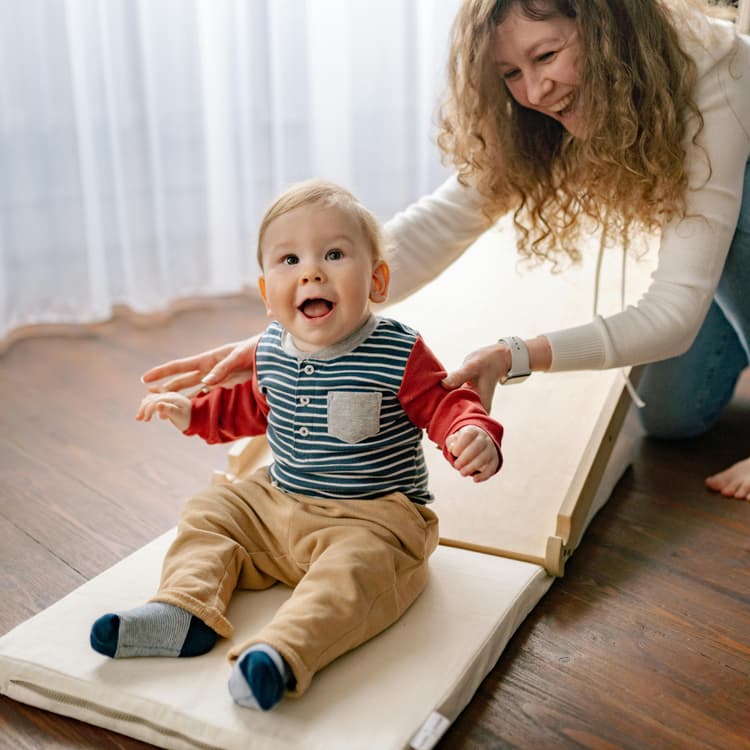 A happy baby down a cushion toboggan with his mother behind him holding him and smiling with him